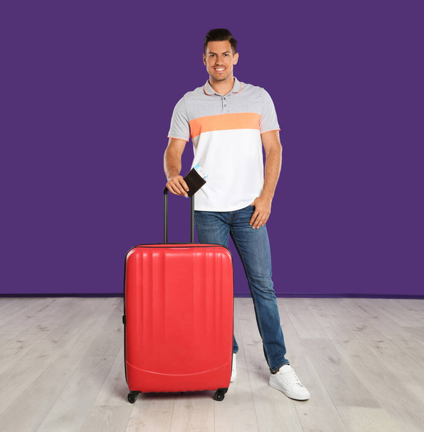Handsome man with suitcase and ticket in passport for summer trip near purple wall. Vacation travel - Fotó, kép