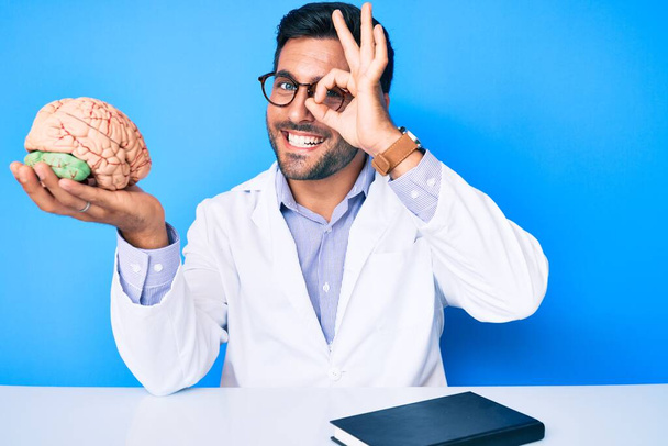 Młody Latynos siedzi przy stole w płaszczu lekarskim trzymając mózg uśmiechnięty szczęśliwy robiąc znak ok z ręką na oku patrząc przez palce  - Zdjęcie, obraz