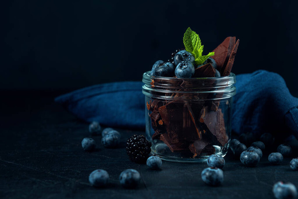 Glass jar with blueberry, blackberry, mint and chocolate pieces on the dark textured background.  - Photo, Image