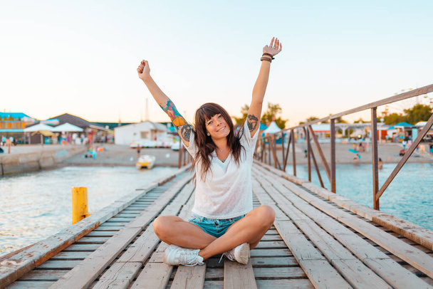 Piękna kobieta z tatuażami siedzi skrzyżowana na drewnianym molo nad morzem, z podniesionymi z radości rękami. Koncepcja wakacji letnich. - Zdjęcie, obraz