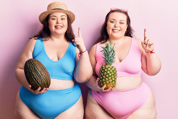 Jóvenes gemelos de talla grande con bikini sosteniendo melón y piña sonriendo con una idea o pregunta apuntando con el dedo con la cara feliz, número uno  - Foto, Imagen