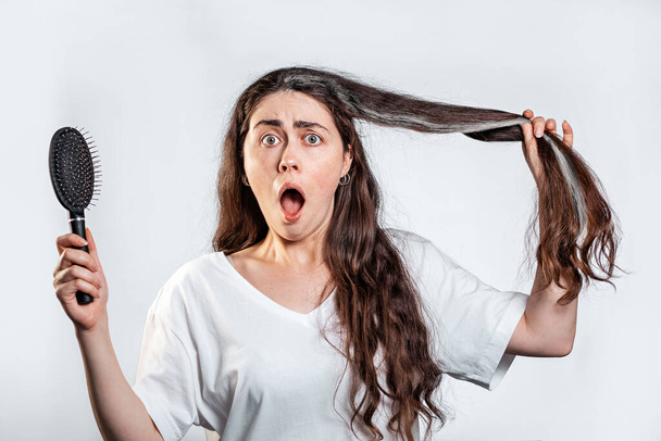 A shocked brunette woman holds a comb and shows a lock of gray hair. White background. The concept of hair loss and gray hair. - Photo, Image