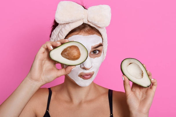 Beautiful spa woman with facial mask on face and holding halves of avocado in hands, looking at camera, doing cosmetology procedures at home, wearing hair band with bow. - Фото, изображение