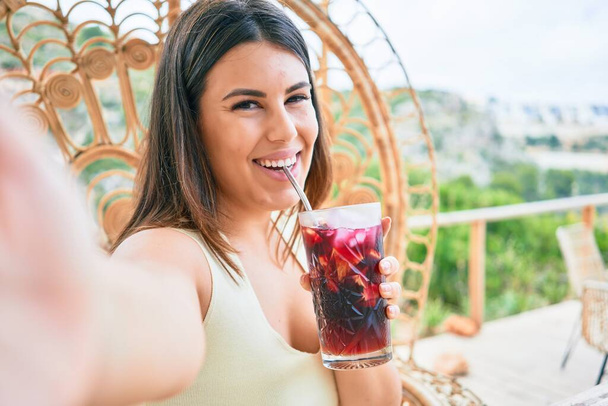 Young beautiful woman smiling happy. Sitting on the table drinking cocktail making selfie by the camera at terrace bar - 写真・画像