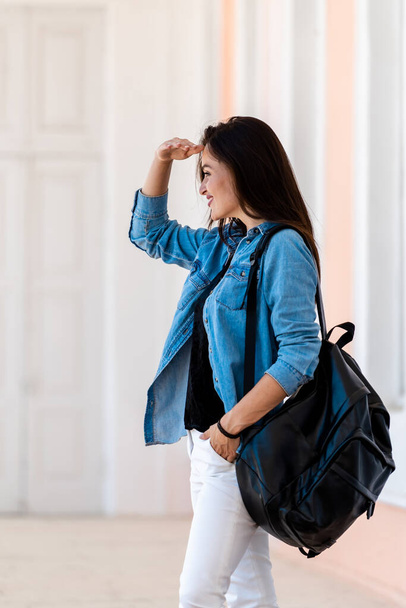 Pretty woman with backpack. View of young woman dressed in blue shirt and white jeans. Looking at side with hand near eyes. View from the side. Half-face view. - 写真・画像