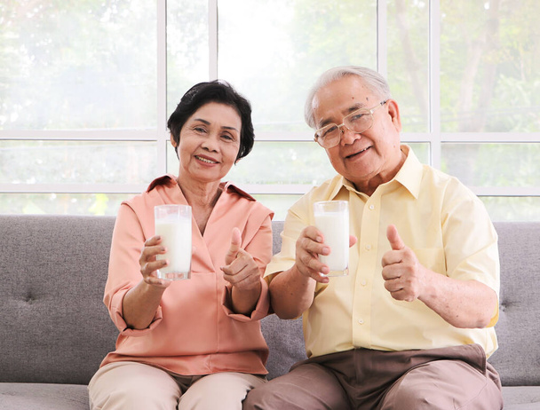 Conceito de estilo de vida de idosos saudáveis. Saudável casal asiático idoso sentado no sofá na sala de estar, segurando óculos de leite mostrando para câmera e mostrando os polegares para cima. - Foto, Imagem