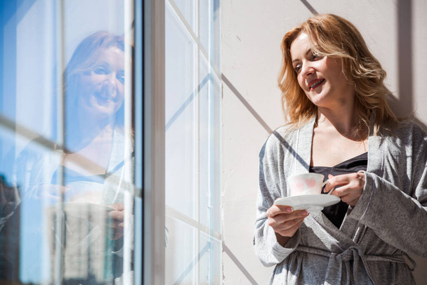 Жінка в срібному одязі стоїть біля вікна з чашкою напою в руках. Погода хороша зовні, сонце сяє. Горизонтальна фотографія
 - Фото, зображення