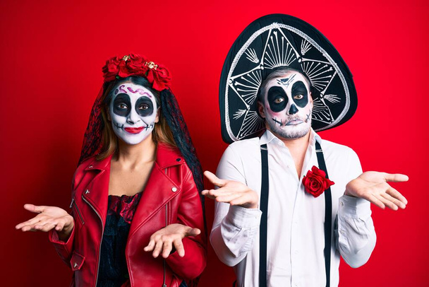 Couple wearing day of the dead costume over red clueless and confused expression with arms and hands raised. doubt concept.  - Photo, Image
