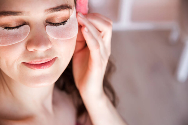Beautiful woman is wearing pink headband rests on a bed early in the morning and put patches under my eyes as a home care. Horizontal photo - Foto, Imagen