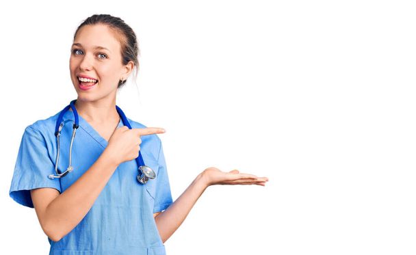 Young beautiful blonde woman wearing doctor uniform and stethoscope amazed and smiling to the camera while presenting with hand and pointing with finger.  - Foto, Imagem