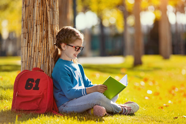 木の近くの芝生の上に座って、同等の宿題をしながらメモ帳で笑顔と読書ノートで幸せな女の子の側面図 - 写真・画像