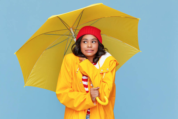 Jovem mulher étnica com guarda-chuva olhando para longe e tremendo no frio dia de outono no backgroun azul - Foto, Imagem