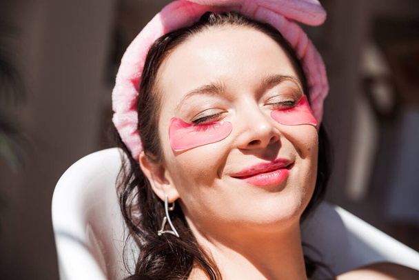Beautiful young woman in a pink cosmetic bandage and with pink birds under her eyes poses in front of the camera in a white ceramic bath in a sunny bathroom. Girl lies with closed eyes. Horizontal photo - Foto, Imagem