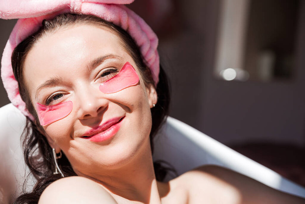 Een mooie jonge vrouw in een roze cosmetisch verband en met roze vogels onder haar ogen poseert voor de camera in een wit keramisch bad in een zonnige badkamer. Meisje lachend en kijkend naar de camera. Horizontale foto - Foto, afbeelding