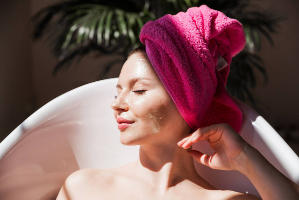 A beautiful young woman with a pink towel on her head and with a golden mask on her face poses for the camera in a white ceramic bathtub in a sunny bathroom. Girl smiles and lies with closed eyes. Horizontal photo - Photo, Image