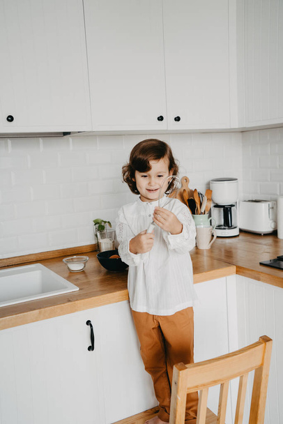 Маленькая девочка готовит тесто для блинчиков на кухне. Концепция приготовления пищи, белая кухня на заднем плане. Серия фотографий повседневного образа жизни в реальном интерьере - Фото, изображение