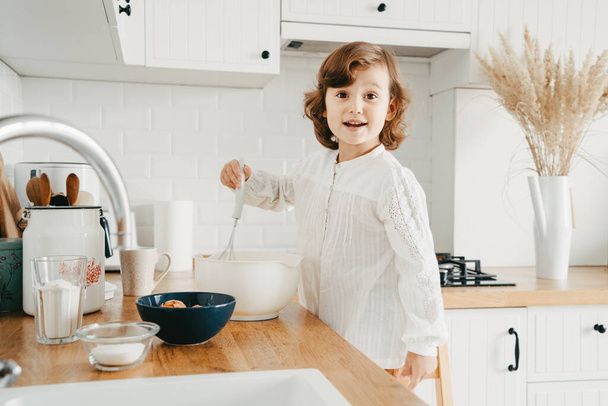 Little girl preparing dough for pancakes at the kitchen. Concept of food preparation, white kitchen on background. Casual lifestyle photo series in real life interior - Φωτογραφία, εικόνα