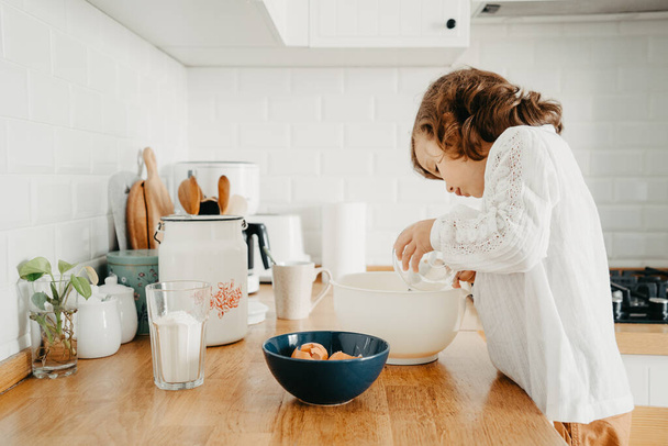 Little girl preparing dough for pancakes at the kitchen. Concept of food preparation, white kitchen on background. Casual lifestyle photo series in real life interior - Fotó, kép