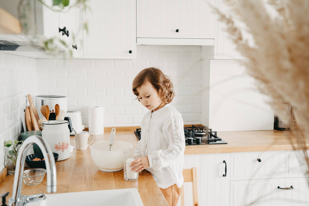Little girl preparing dough for pancakes at the kitchen. Concept of food preparation, white kitchen on background. Casual lifestyle photo series in real life interior - Фото, изображение