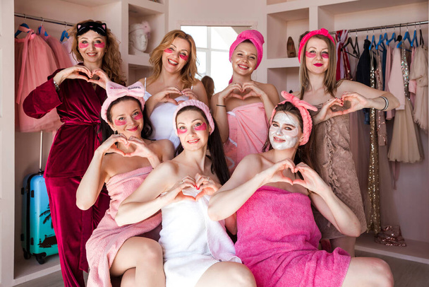 Siedem pięknych młodych kobiet w różowych ręcznikach, z kosmetycznymi bandażami na głowach, pozuje do aparatu z łatami pod oczami i maską kremową na twarzy. Koncepcja opieki domowej. Dziewczyny z palcami pokazują znak serca, symbol miłości. Poziomy - Zdjęcie, obraz