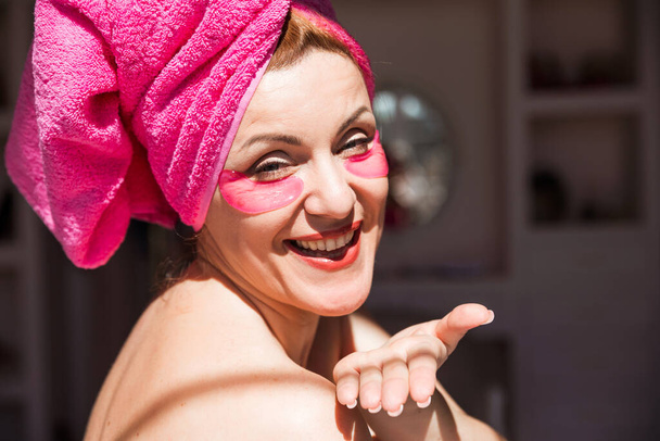 Schöne Frau mit einem rosa Handtuch auf dem Kopf und mit rosa Flecken unter den Augen schickt einen Luftkuss in die Kamera. Horizontales Foto - Foto, Bild
