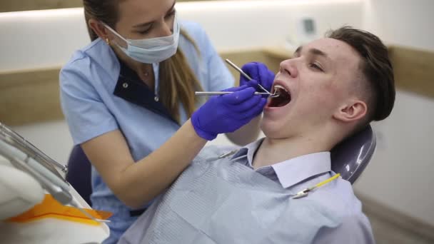 Οδοντίατρος ορθοδοντική γυναίκα εξετάζει τα δόντια νεαρός άνδρας του ασθενούς χρησιμοποιώντας οδοντικά εργαλεία βελόνα και καθρέφτη. Στοματική υγιεινή και θεραπεία. - Πλάνα, βίντεο
