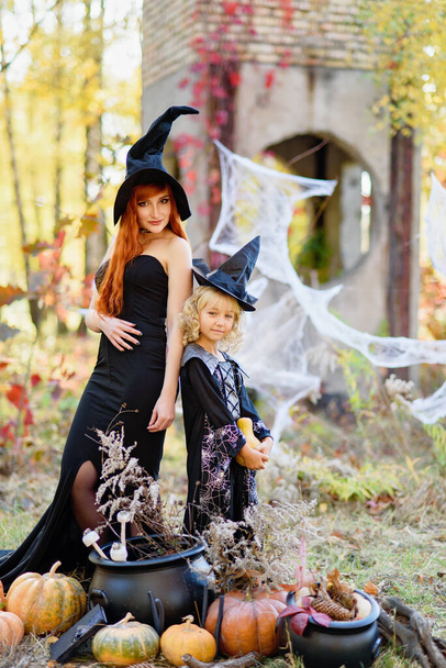 Dziewczynka i kobieta w stroju czarownicy świętują Halloween na świeżym powietrzu i baw się dobrze. Cukierek albo psikus. Czarownica z magiczną Księgą zaklęć. Tło Halloween. Latarnia dyniowa z głowicą dyni - Zdjęcie, obraz