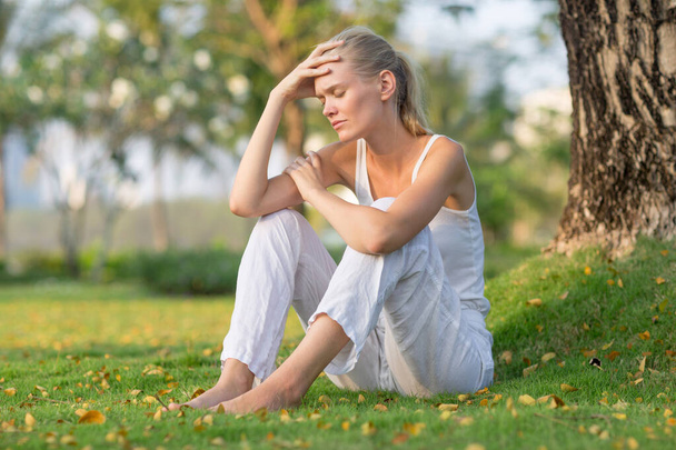 Smutná žena sedící sama na trávě v parku s rukou na čele. Špatný pocit a deprese. - Fotografie, Obrázek