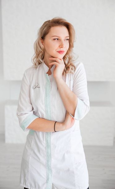 Krásná mladá žena lékař v bílé uniformě pózovat proti bílé zdi pozadí a při pohledu na kameru. Svislá fotografie - Fotografie, Obrázek