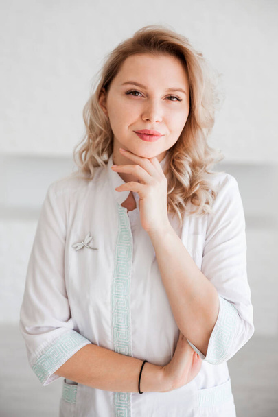 Krásná mladá žena lékař pózuje v bílých lékařských šatech na bílém pozadí zdi. Svislá fotografie - Fotografie, Obrázek