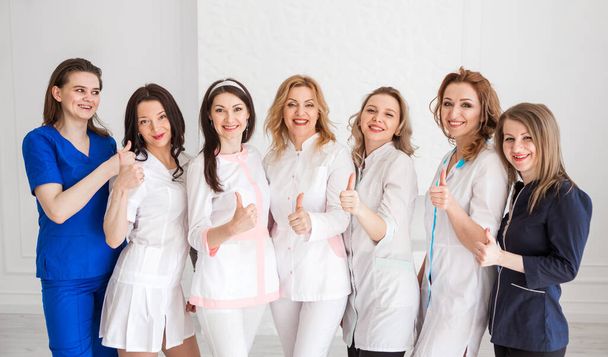 Красивые молодые женщины-врачи в белых мундирах позируют на фоне белой стены, показывая класс рукой, смотрят в камеру и улыбаются. Горизонтальное фото - Фото, изображение