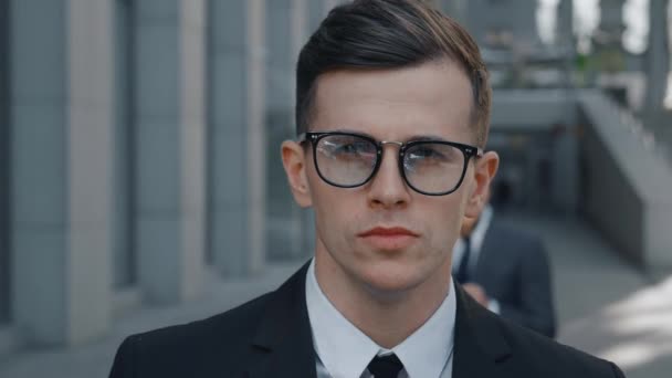 Portrait of attractive business man in suit and eyeglasses looking into camera and outdoors at city downtown. Portrait of handsome male businessman standing outdoor near office building - Footage, Video
