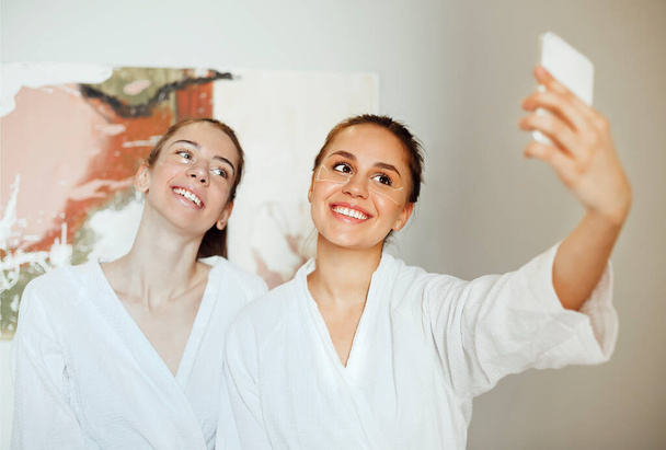Μια παρέα από χαρούμενες φίλες με μπουρνούζια και με μπαλώματα ματιών που βγάζουν selfie στο smartphone ενώ χαλαρώνουν στο spa center το Σαββατοκύριακο - Φωτογραφία, εικόνα