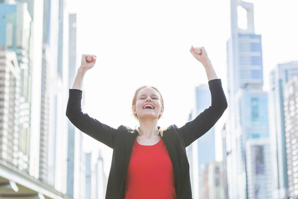 Діловий та кар'єрний успіх - Щаслива молода бізнес-леді святкує робочі досягнення обома руками на фоні висотних будівель сучасного міста
. - Фото, зображення