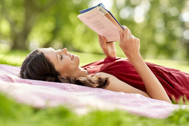 gelukkig vrouw lezen boek liggend op deken in park - Foto, afbeelding