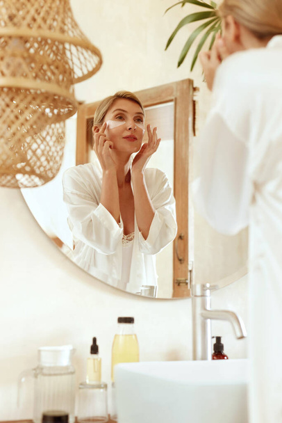 Skincare Routine. Woman With Patches Under Eyes Standing In Bathroom And Looking At Mirror. Lady In Fashion Silk Pajama Using Hydrogel With Hyaluronic Acid And Collagen For Beauty Procedure At Home. - Фото, зображення