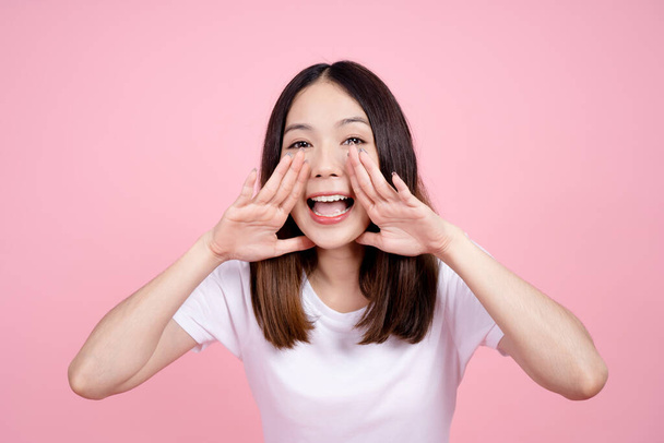Beautiful Asian woman shouting and looking up, using her mouth to tell something or call someone on a pink background. - Photo, Image