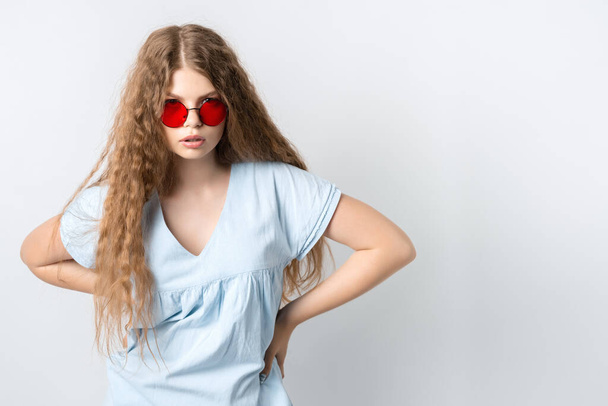 Portret charyzmatycznej nowoczesnej dziewczyny z kręconymi długimi włosami w lekkich ubraniach i nosi okrągłe czerwone okulary, stanowiące izolowane na białym tle ściany. Koncepcja stylu życia ludzi - Zdjęcie, obraz
