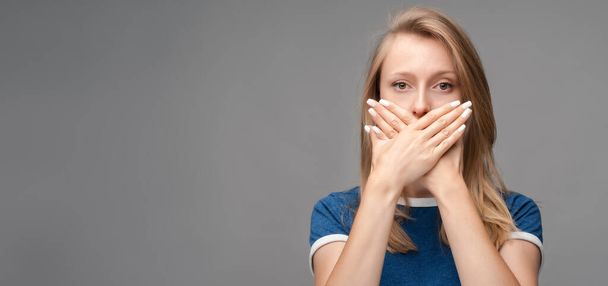 Schockierte junge blonde Frau, gekleidet in blaues T-Shirt, den Mund mit den Händen auf grauem Hintergrund verdeckt. Behalten Sie Schweigen Konzept. Kopieren Sie Platz für Ihren Text - Foto, Bild