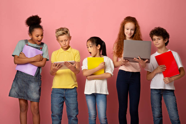 Portrait of multiethnic schoolchildren with notebooks using tablet and laptop to study on pink background - Photo, Image