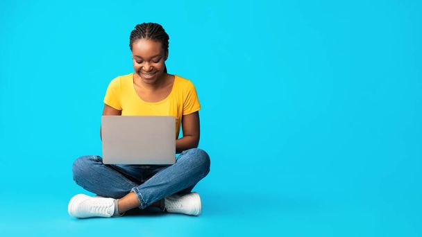 青い背景の上に座ってノートパソコンを使用して幸せな黒の女の子,パノラマ - 写真・画像
