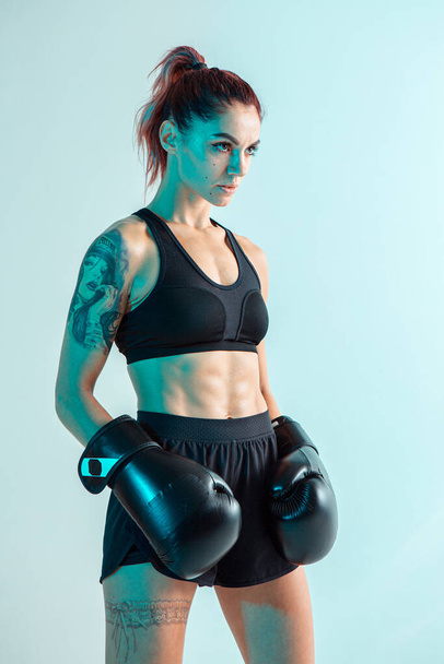 Atletica combattente ragazza in guanti da boxe in posa in studio in luce blu neon. Manifesto di arti marziali miste. Corpo con tatuaggio - Foto, immagini