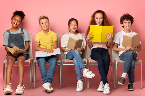 School education. Happy diverse kids with textbooks and notebooks sitting on chairs against pink background - Foto, imagen