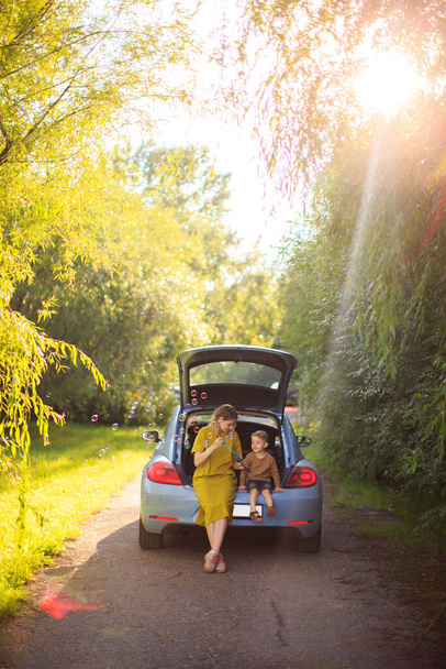 Beautiful mother and baby boy sit in trunk of car on road and blow soap bubbles. Summertime photo about family travel - Photo, Image