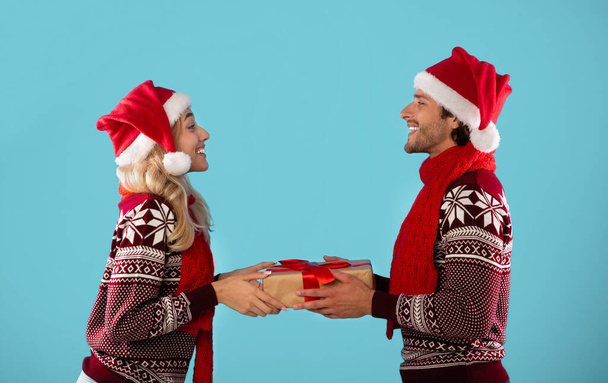 Side view of millennial couple in warm sweaters and Santa hats holding Christmas gift on blue background - Zdjęcie, obraz