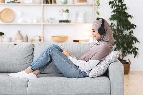 Weekendowa rozrywka. Arabski dziewczyna relaks w domu ze smartfonem i słuchawki bezprzewodowe - Zdjęcie, obraz