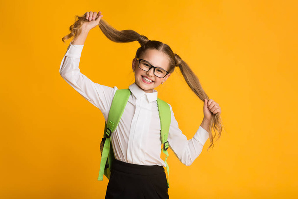 Pequena estudante brincando com o cabelo posando no fundo amarelo - Foto, Imagem