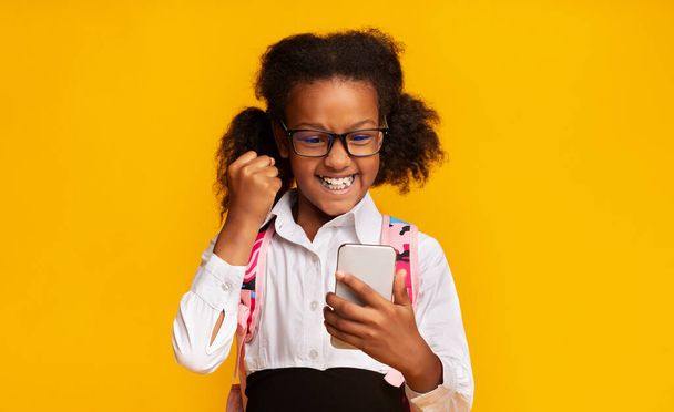 Elementary Student Girl With Smartphone Shaking Fists, Yellow Background - Foto, Imagen