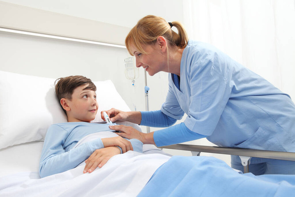 Happy child lying in bed in hospital room and nurse taking his temperature  - Photo, Image
