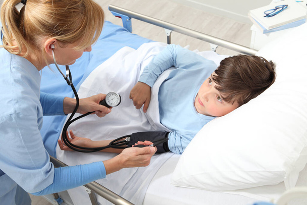 Дитина лежить в ліжку в лікарняній кімнаті і медсестра вимірює його тиск за допомогою сфінгометру і стетоскопа
 - Фото, зображення